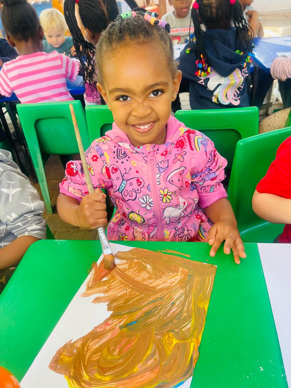 A girl in a pink jacket smiling while painting at the craft table